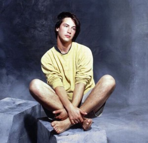 A twentysomething year old Keanu Reeves sits cross-legged on a grey block in front of a grey photographer’s dropcloth. He’s wearing khaki shorts and a yellow sweatshirt with the sleeves pushed up. He stares off into the distance with his surfer-styled hair swept to one side.