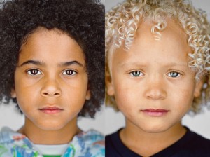 An image of two young mixed race boys. One has a head of tight black curls and dark eyebrows. The other has has light blond, tightly curled hair and light eyebrows and eyes.  