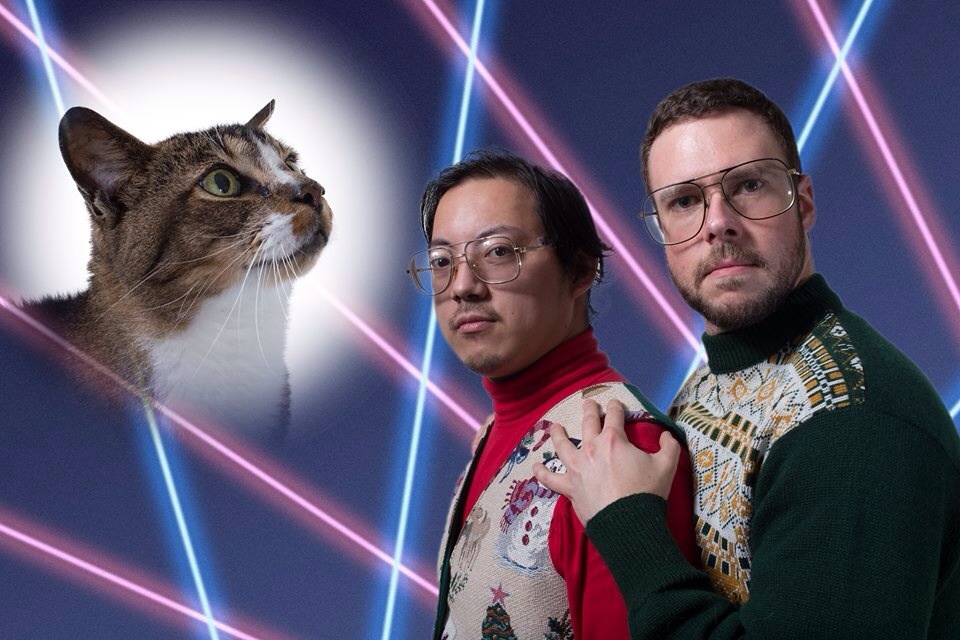 A white man and an Asian man, both wearing ugly Christmas sweaters and aviator style eyeglasses, pose against a backdrop of lasers, like the old school photo trend in the 80s, with an inset of a cat.
