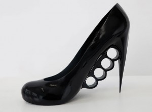 A black high-heeled women's shoe where the heel is a very sharp point and the sole has finger holes like a set of brass knuckles.