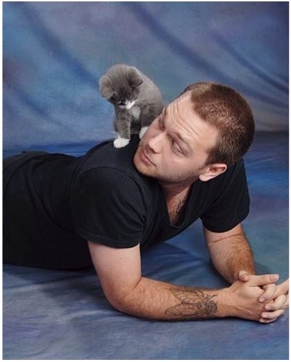 A white man in a black t-shirt lies on the floor, propped up by his forearms and looks up at the grey and white kitten on his shoulder.