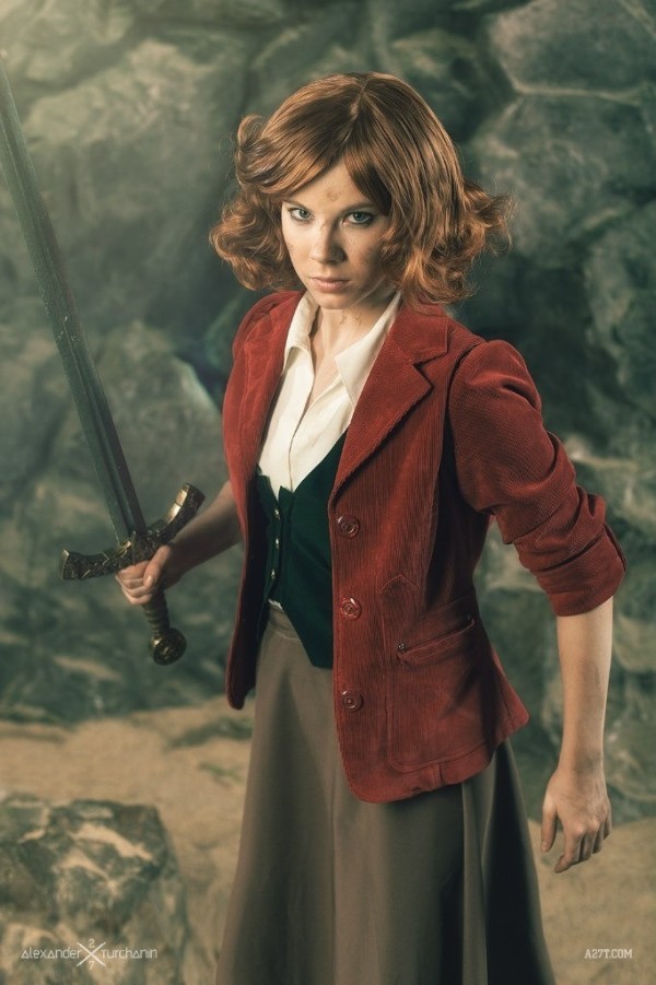 A white woman with red hair holds a sword in her right hand and is dressed in a white shirt, black waistcoat, red blazer and long brown skirt.