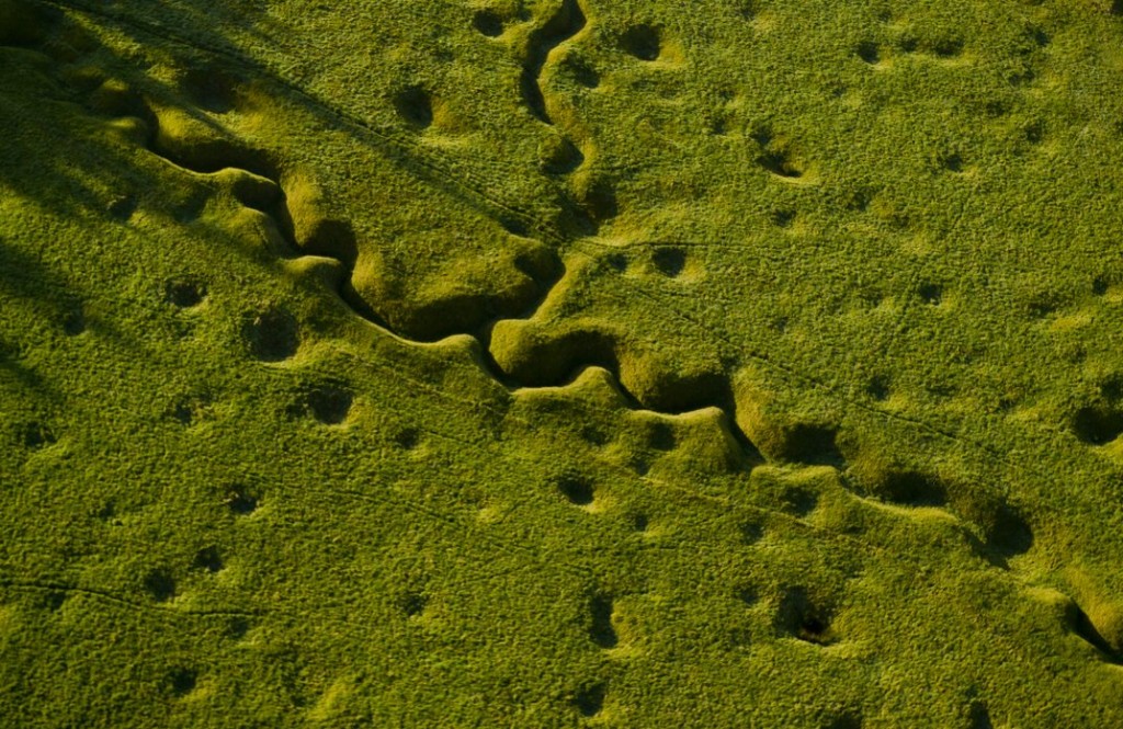 An aerial photo of a WWI battlefield today. It's covered in greenery but still looks pockmarked from the bomb craters and shows a long, snaking trench across the middle of it.