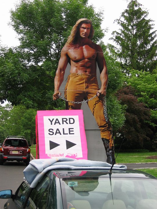 A large standee of a shirtless Fabio wearing tan suede pants with his trademark long blond hair, sits on top of a car holding a hot pink sign advertising a yard sale. 