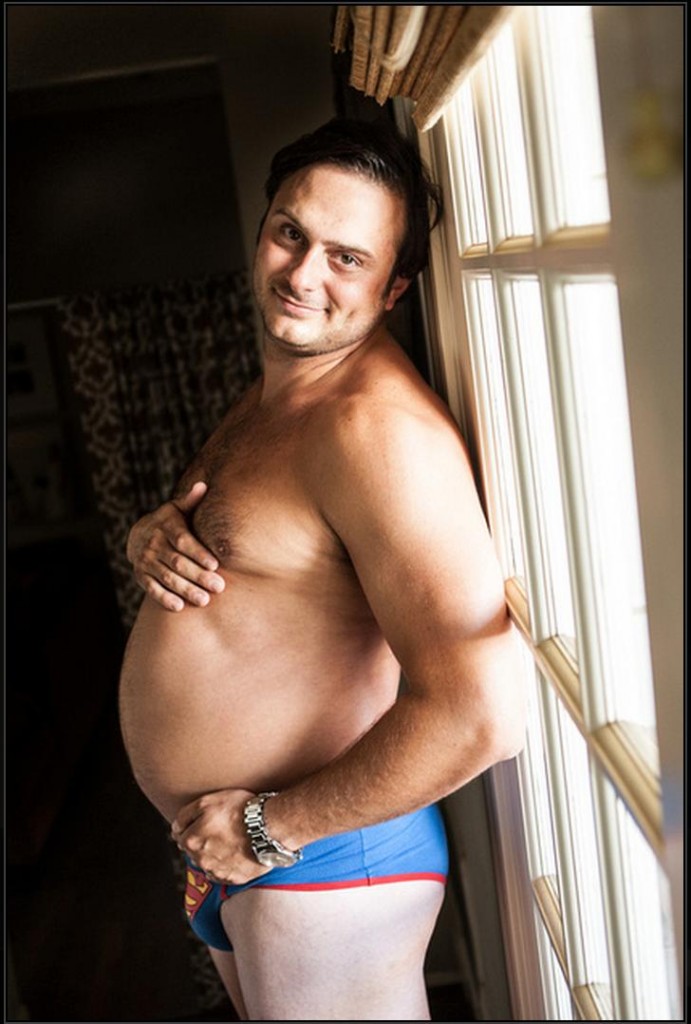 A white man, wearing only a pair of blue and red Superman briefs, frames his beer belly with his hands and smiles at the camera in the style of maternity portraiture.