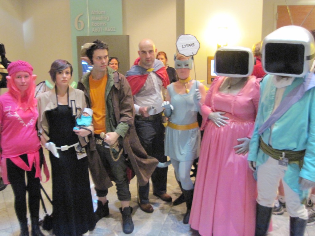 A group of 6 people cosplaying as characters from the comic Saga. The prince and his wife are dressed like 19thc aristos but with tv monitor heads. Lying cat is a white woman in a grey catsuit holding a sign that says "lying." The Will is a bald white man wearing a red cape with dark pants and light shirt. Marko has goat horns and wears an orange shirt with brown pants and trench coat.  Alana is a white woman with  fairy wings who wears a long black dress and holds a baby doll.  Izabel the ghost girl is a white woman in a pink wig and dress and black tights.