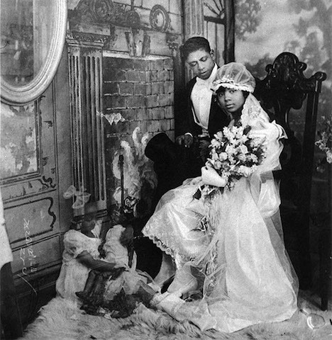 A young black couple poses for a wedding photo in the 20s or 30s. He wears a dark tux with a white shirt and she wears a tea length white gown with a full length lace veil and sits in a chair holding a large bouquet.  At their feet is a ghost image of young children suggesting the future children of a happily married couple.