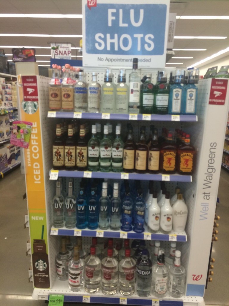 A photo taken inside a Walgreens shows shelves stocked with various brands of liquor. The sign says FLU SHOTS.