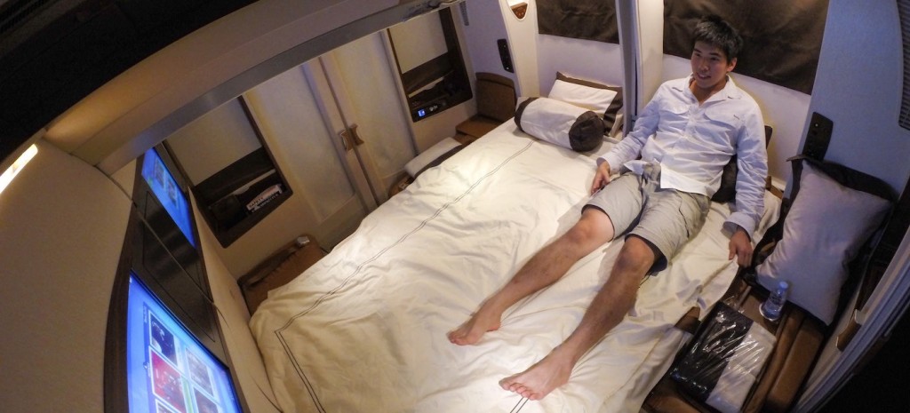 An Asian man in khaki shorts and a blue oxford shirt sits comfortably and watches tv on a double bed during a flight from Singapore to NYC.