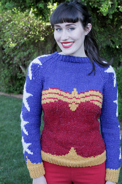 A white woman with dark brown hair, pale skin and bright red lips smiles as she models a sweater in a lush backyard.  The body of the sweater is red and gold in the outline of Wonder Woman's corset top. The chest and sleeves are blue, with white stars running from collar to sleeve cuff. The cuffs are gold. 