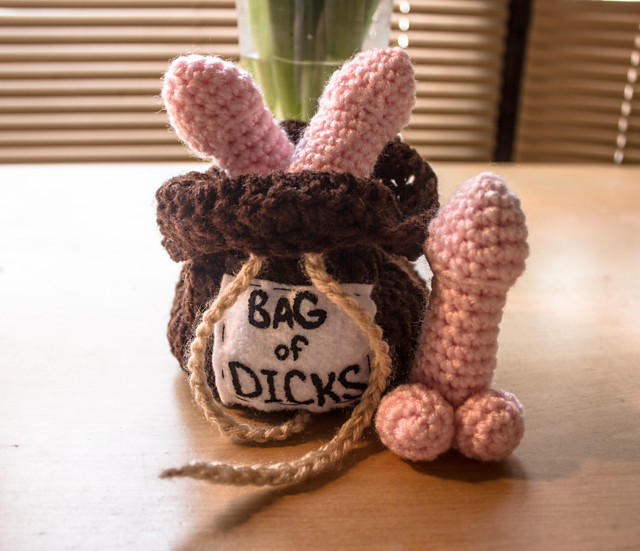 crocheted pink phalluses peek out of a brown crocheted bag with a label on it that says "BAG of DICKS"