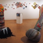 An art gallery with white walls and a shiny wood floor. 5 foot tall phalluses made from colorful strips of fabric sewn together in random intervals stand around the room.