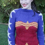 A white woman with dark brown hair, pale skin and bright red lips smiles as she models a sweater in a lush backyard. The body of the sweater is red and gold in the outline of Wonder Woman's corset top. The chest and sleeves are blue, with white stars running from collar to sleeve cuff. The cuffs are gold.