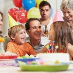 A stock photo of a child's birthday party. A white father is flanked by a young son and daughter at a table with a birthday cake on it. A white mother and grandmother look on. In the back, with the balloons, stands a middle-aged white man in a white tank top holding a beer and looking out at the viewer.