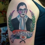 A color tattoo of Ruth Bader Ginsburg from the chest up in her black robe and distinctive lace collar. Laurel frames the sides and SUPREME is written on a scroll below.