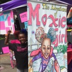 a young black girl with afro puffs stands flexing her biceps next to a tall banner with the illustrated cover for her comic The Adventures of Moxie Girl on it